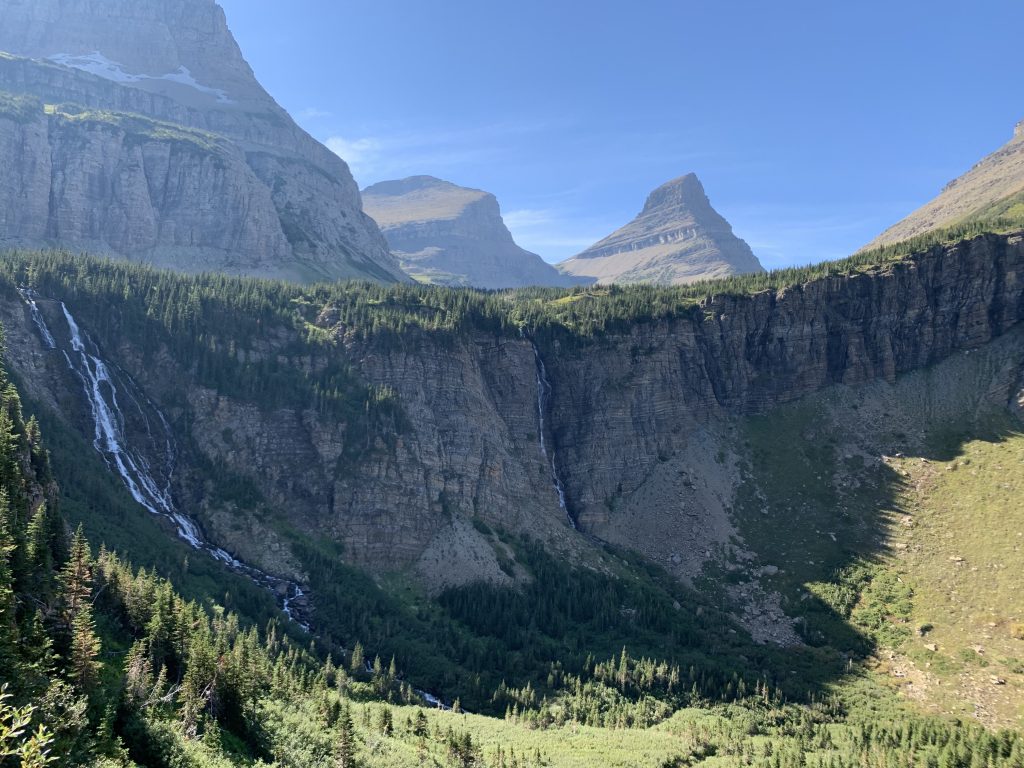 A panoramic view of a steep rocky cliff in Glacier National Park, adorned with waterfalls flowing into a dense forest below, with majestic mountain peaks rising in the background under a clear blue sky