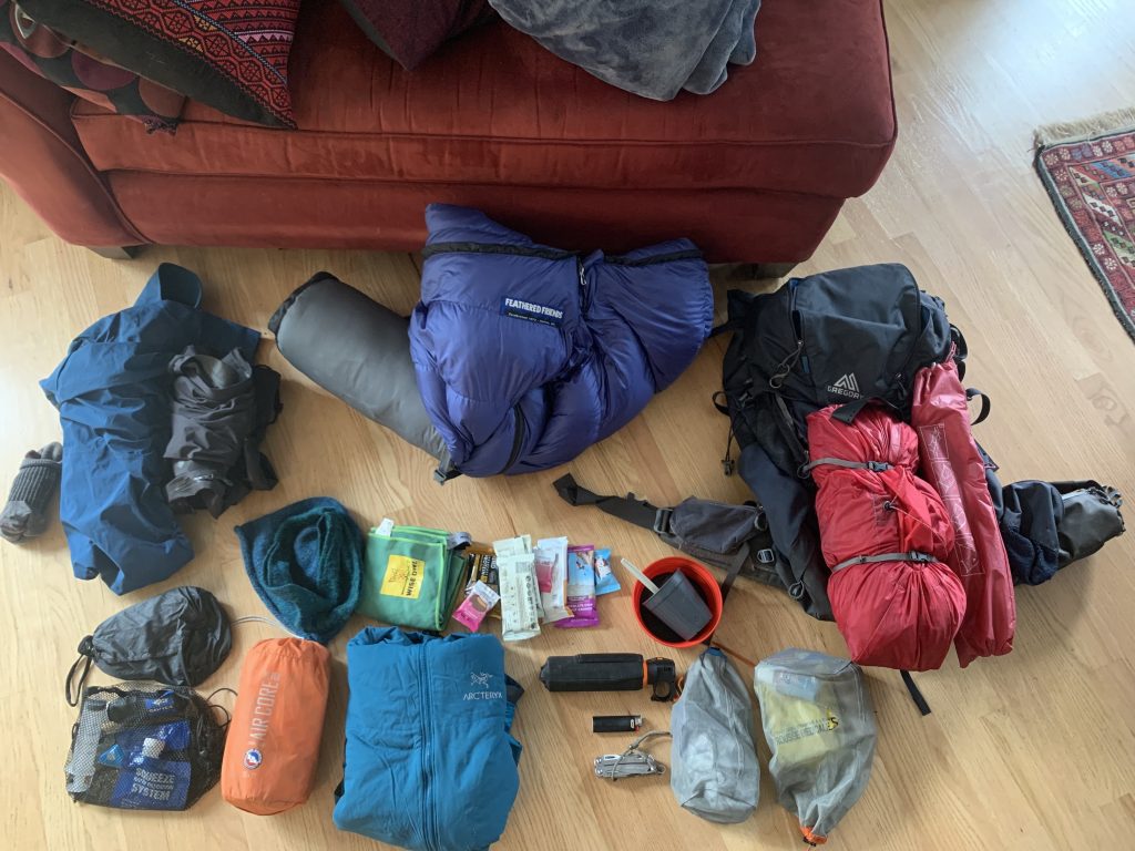 A collection of backpacking gear laid out on the floor, including a blue Feathered Friends sleeping bag, a Gregory backpack, and various clothing, snacks, and camping essentials