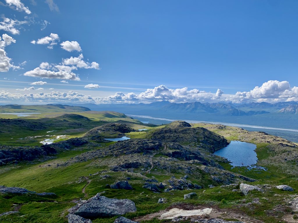 Rolling green hills under a bright blue sky with fluffy clouds, with distant mountains and lakes visible