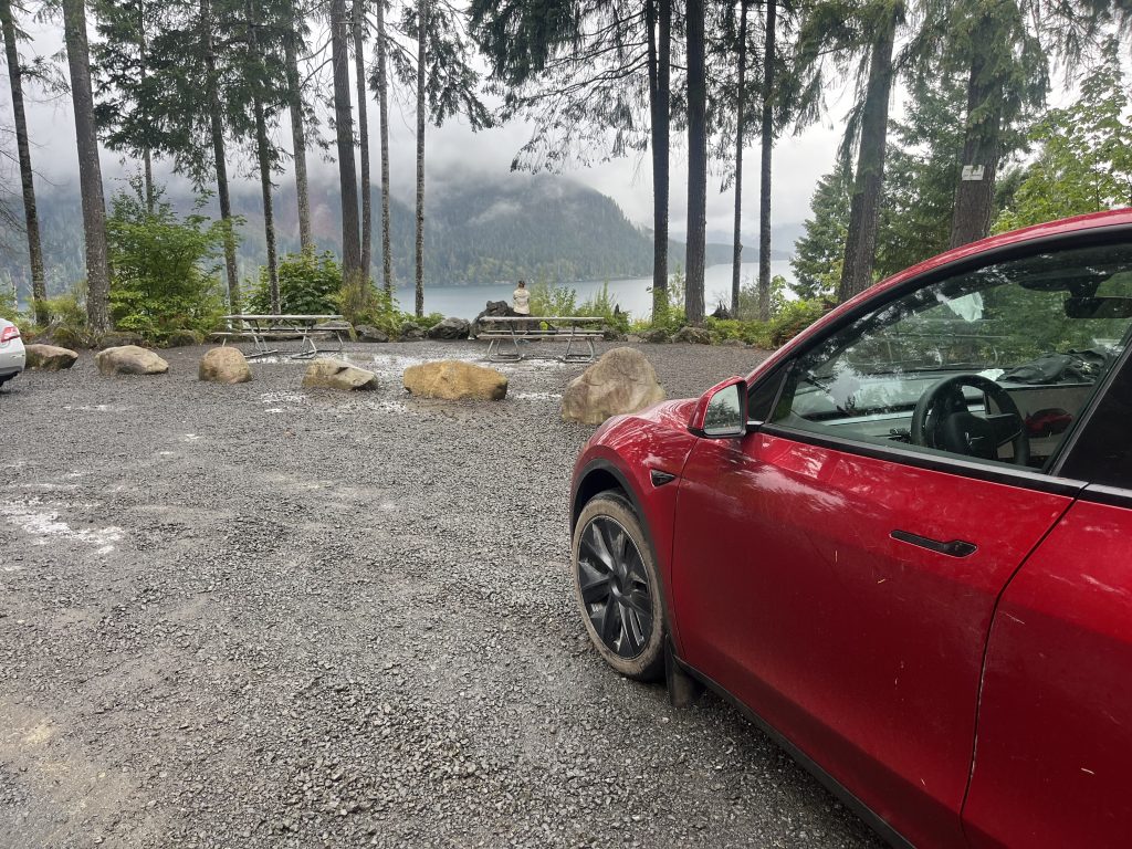 Red Tesla parked at a gravel campsite with a forest and a lake in the background, surrounded by picnic tables and boulders