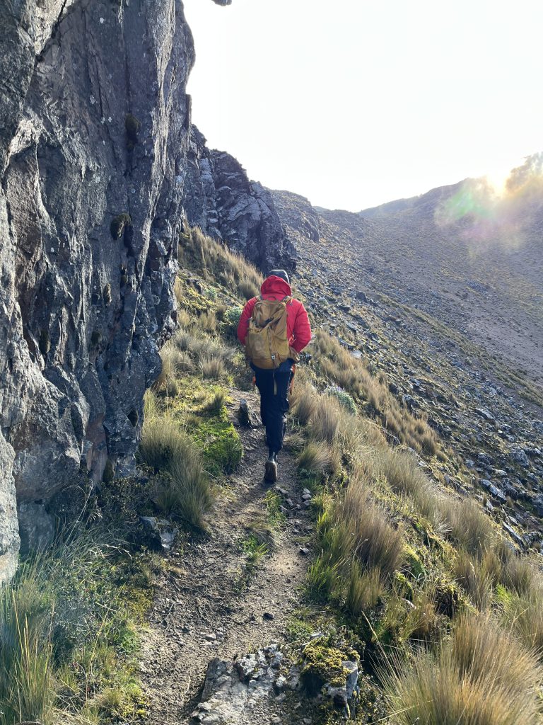 Hiker in a red jacket walking on a rocky trail on a sunlit mountainside