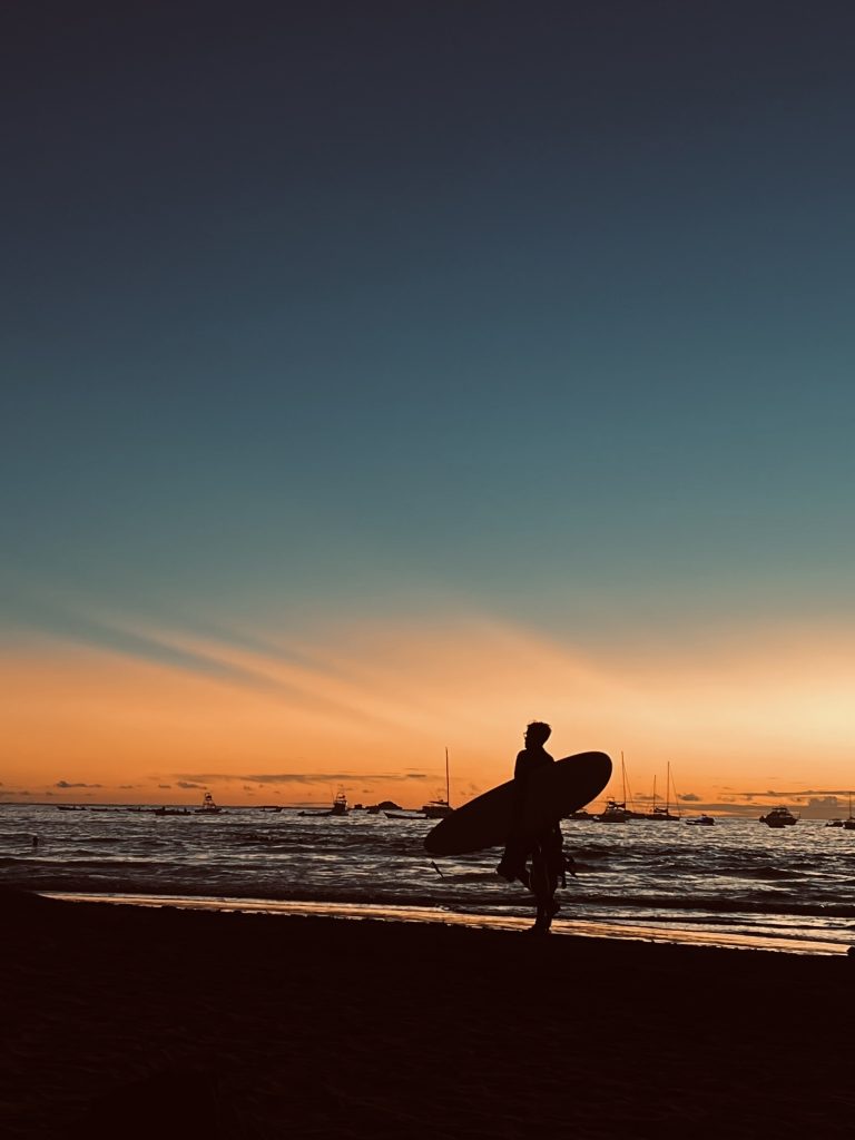 Silhouette of a surfer walking along the beach at sunset in Costa Rica, with boats anchored in the background