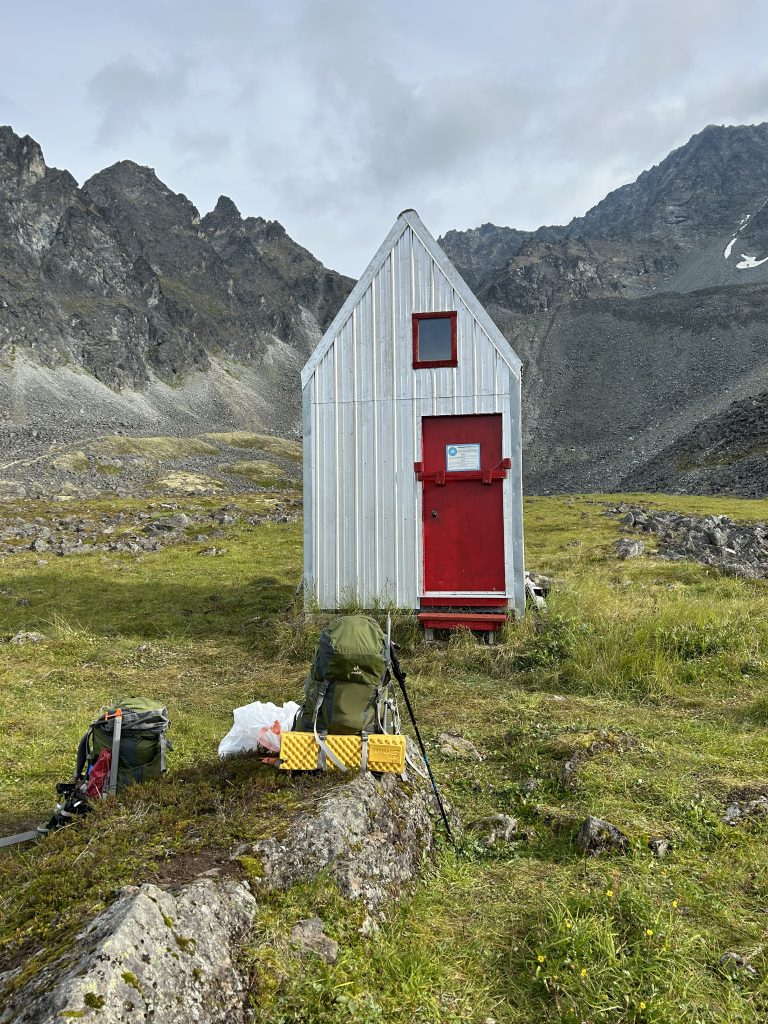 Small silver hut with a red door in a mountainous alpine setting, with backpacks and camping gear placed outside