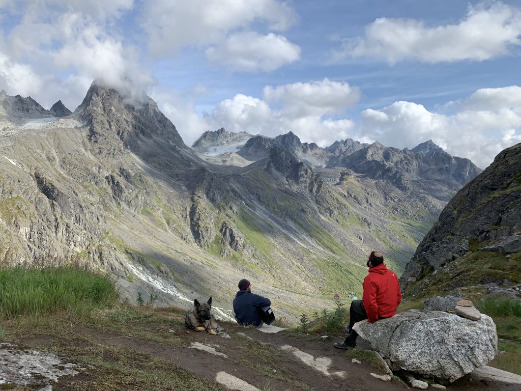 Two people sitting on a rock outcrop with a dog, enjoying a scenic mountain view