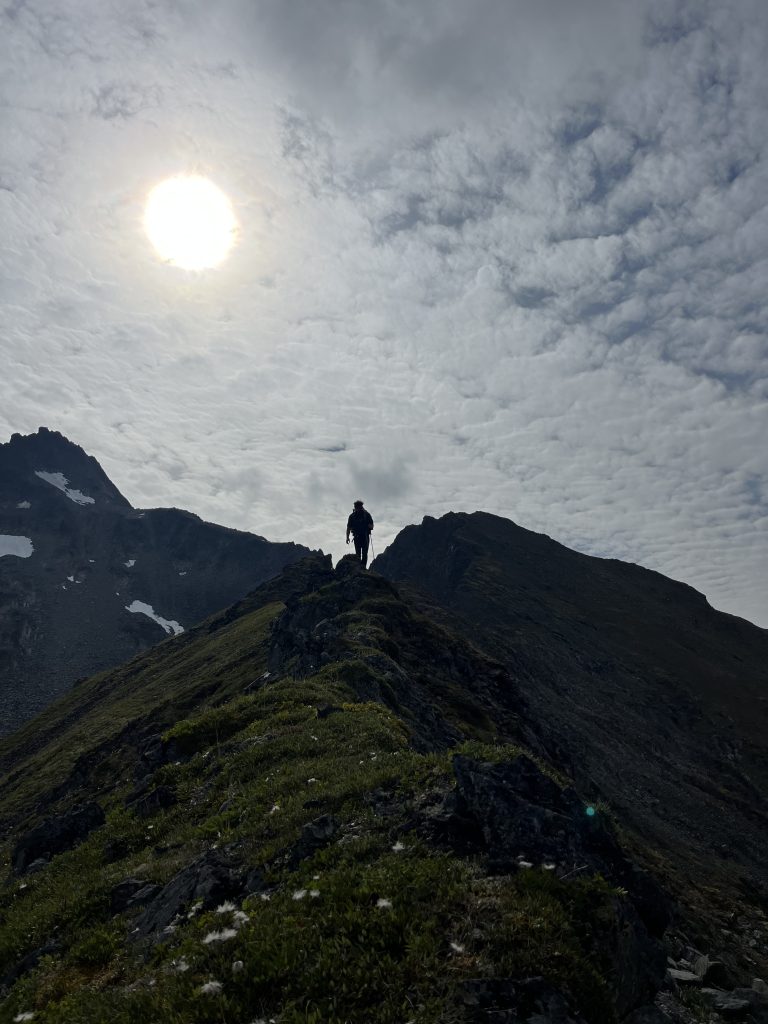 A lone hiker silhouetted against the sun as they stand on a narrow mountain ridge with a partly cloudy sky above