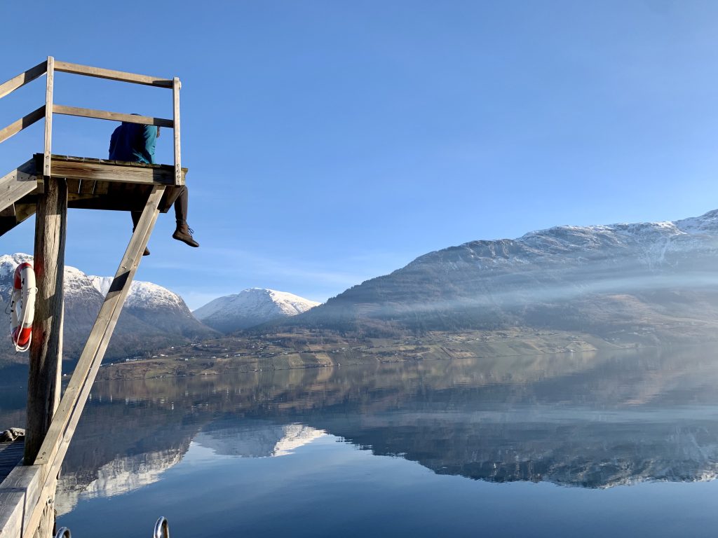 Person sitting on a dock overlooking a calm lake with mountains reflecting in the water