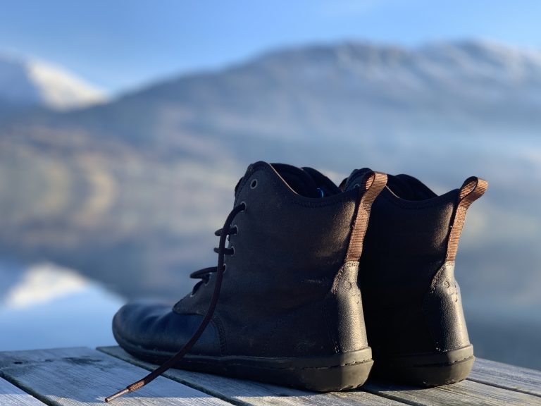 The back of a pair of hiking boots placed on a wooden dock, with a mountain range reflected in the lake behind them