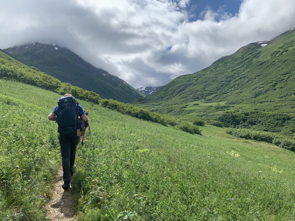 A hiker with a large backpack walking along a green trail in a mountain valley, with cloudy peaks in the background