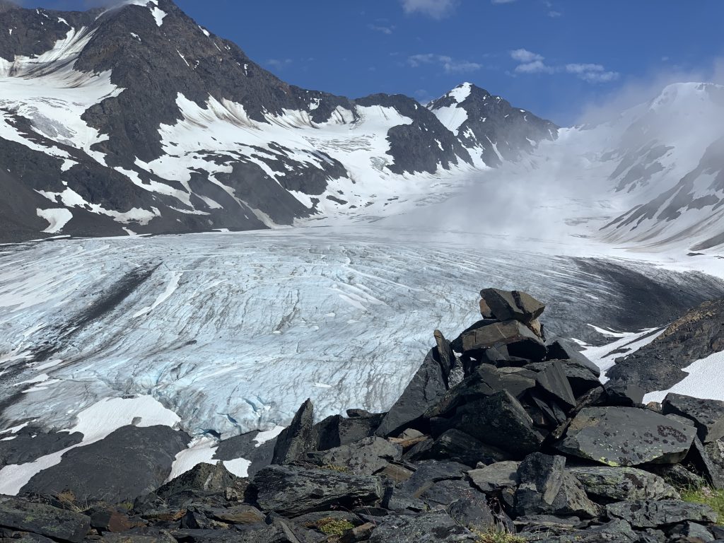 Rocky terrain in front of a massive Raven Glacier at Crow Pass, surrounded by snowy mountain peaks