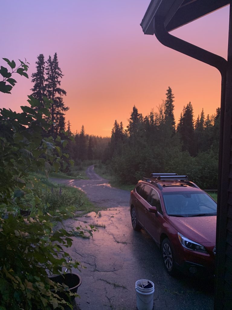 Sunset view from a forested driveway with a red car and golden sky