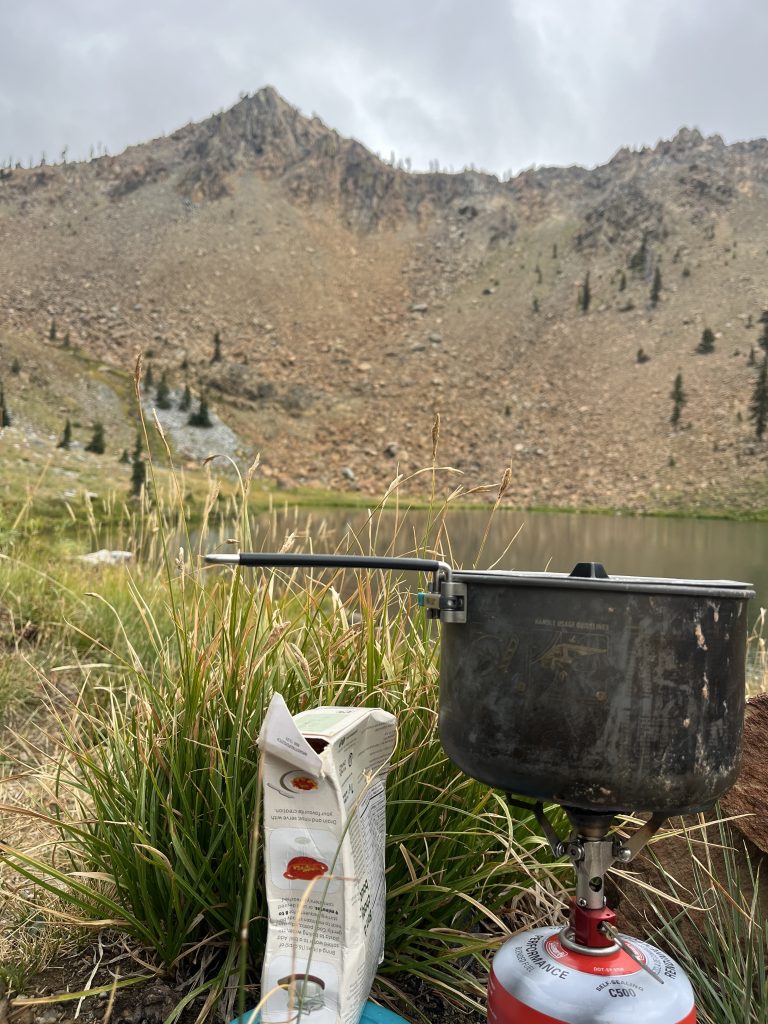 Camping stove with a pot and boxed food near a grassy lakeside with rocky mountains in the background