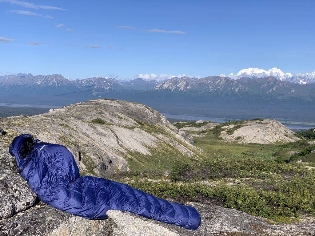 A blue Feathered Friends sleeping bag lying on a rocky outcrop with a breathtaking view of Denali and the Alaskan wilderness under a clear blue sky.
