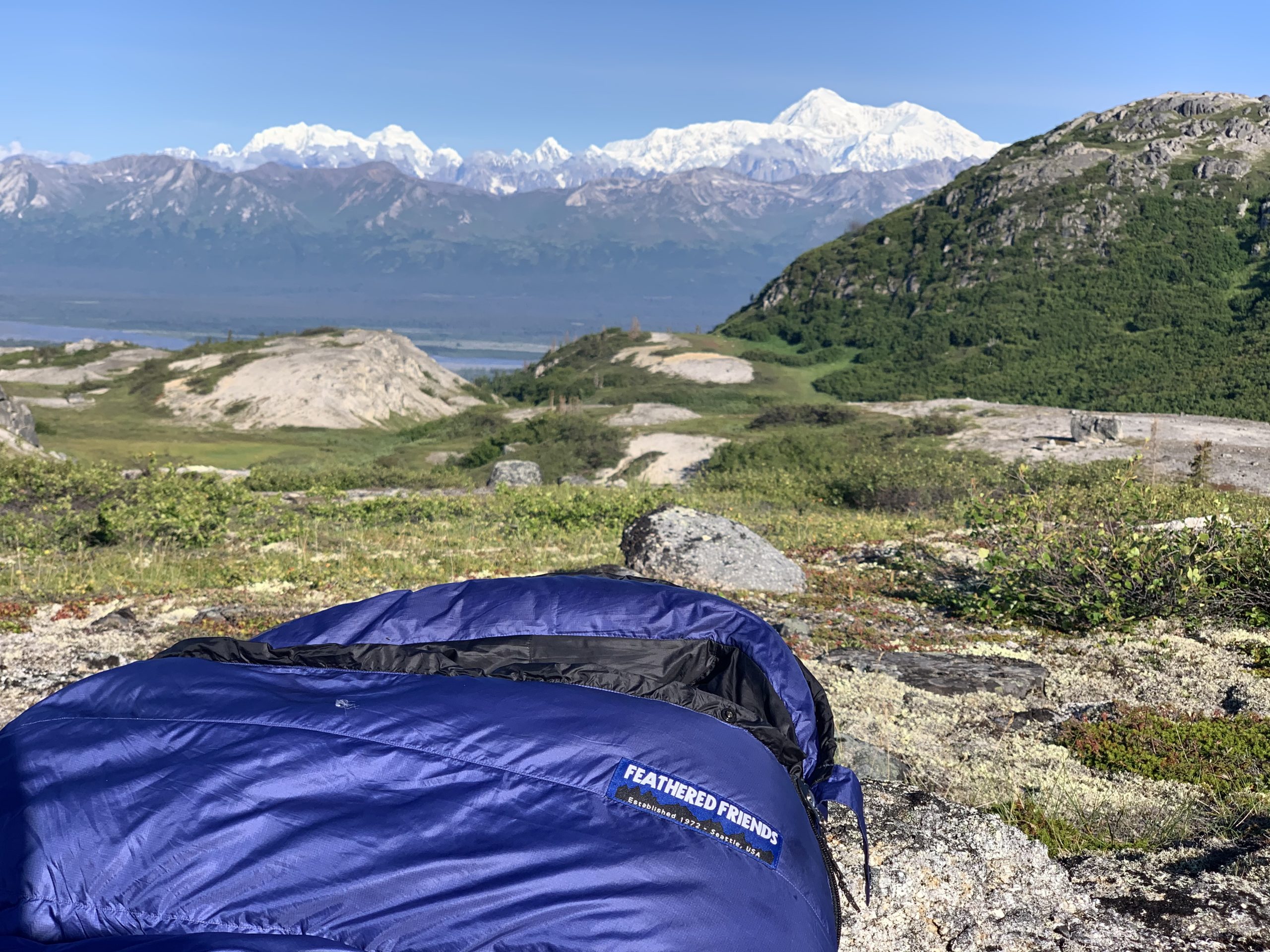 A blue Feathered Friends sleeping bag lies on rocky terrain in the foreground, with a stunning view of mountains, including Denali, under a clear blue sky in the background