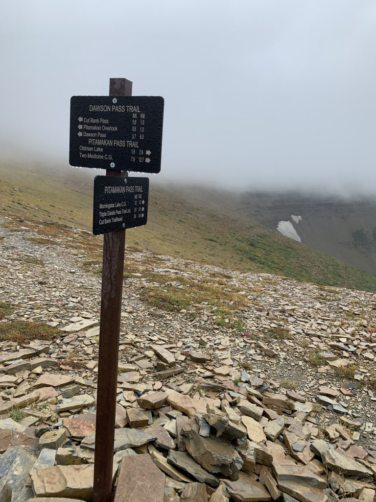 A mountain pass trail sign surrounded by rocky terrain, with thick fog obscuring the background