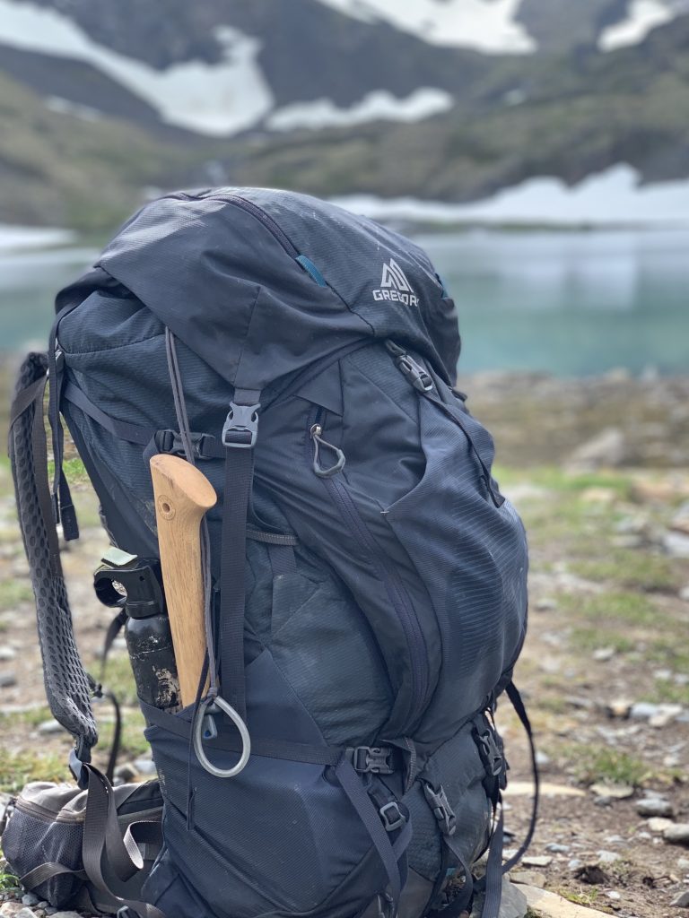 A large Gregory backpack with an axe strapped to its side, standing next to a mountain lake with snowy peaks in the distance.