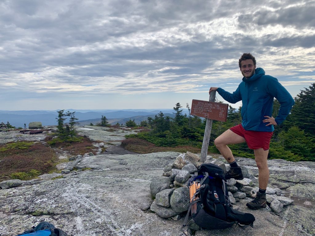 A hiker standing next to a summit sign on a rocky landscape, with cloudy skies and distant mountain ranges in the background.