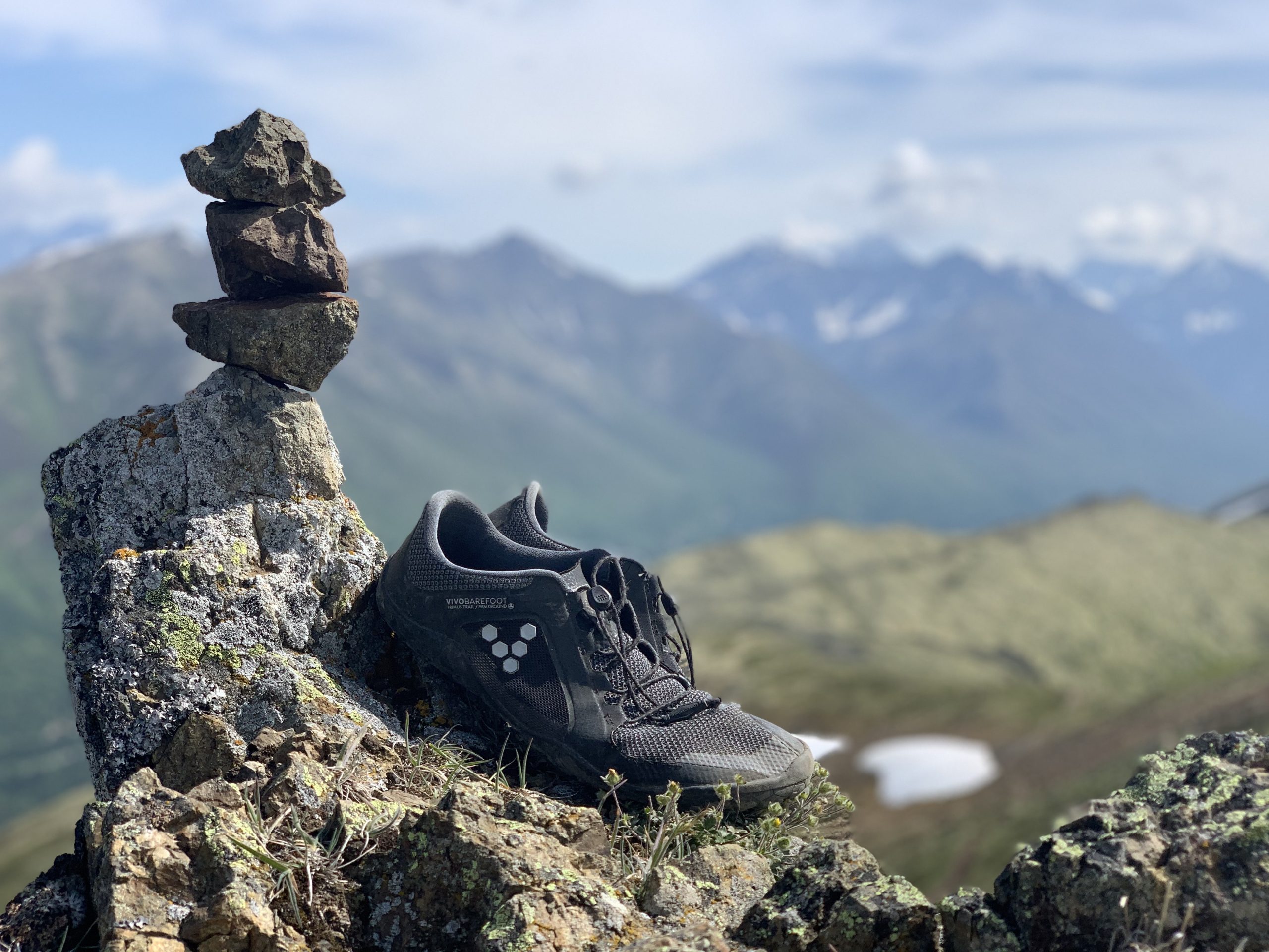 A pair of minimalist hiking shoes next to a stack of balanced rocks on a mountaintop, with a mountain range in the background