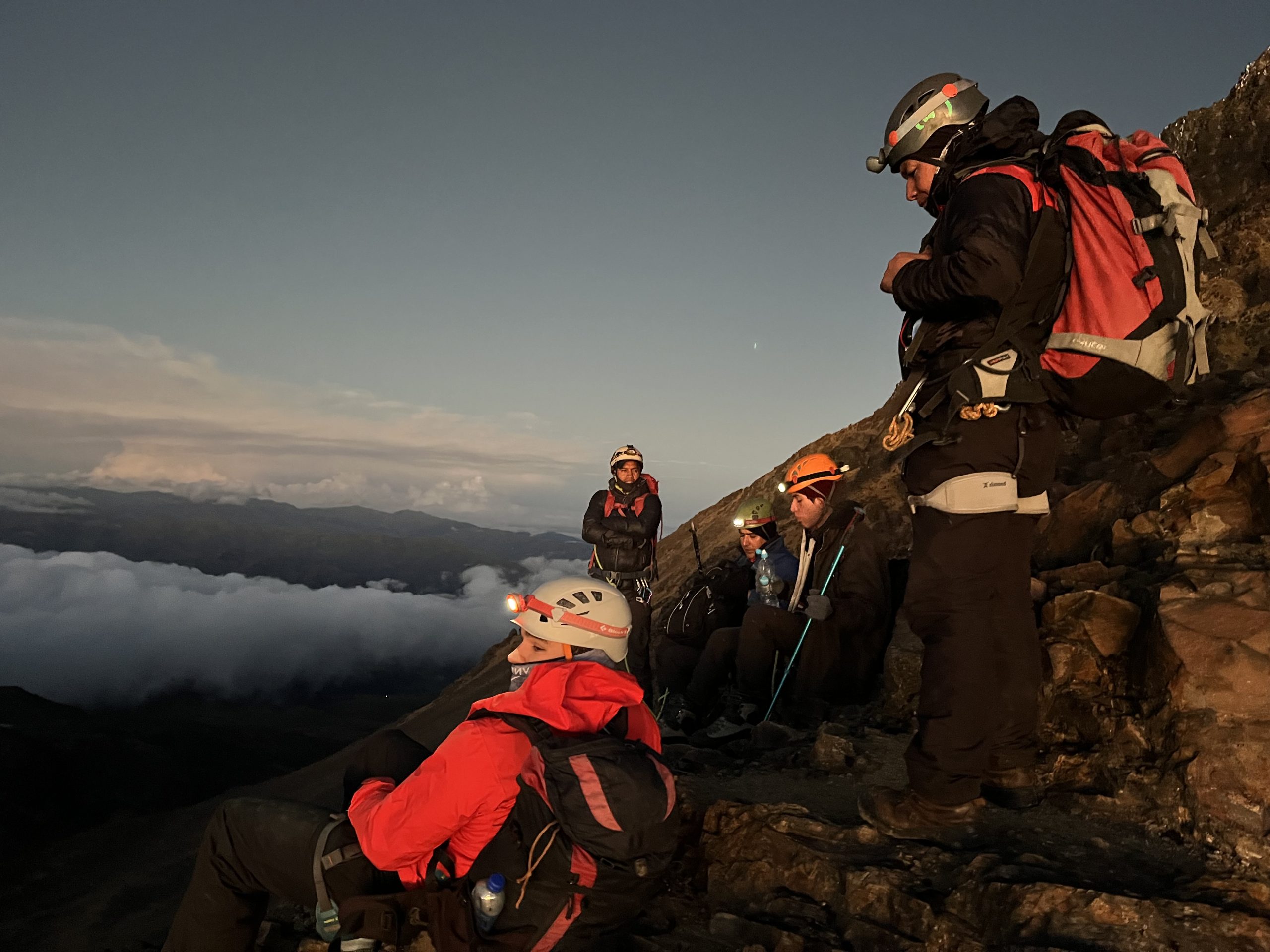 A group of mountaineers wearing helmets and backpacks resting on a rocky ledge during twilight, with clouds in the background