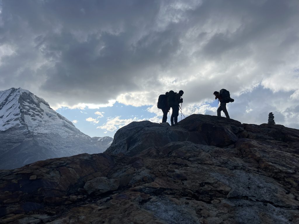 Three hikers with backpacks standing on a rocky mountain ridge, silhouetted against a cloudy sky with snow-covered mountains in the background