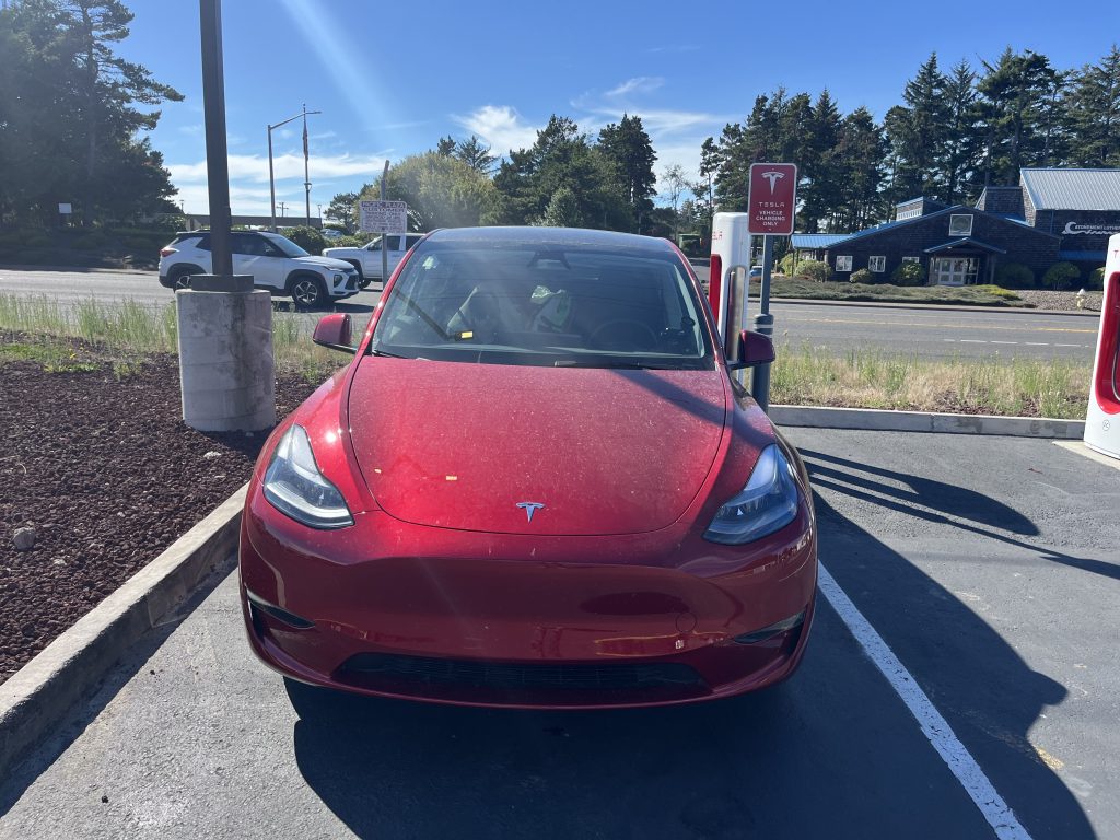 Red Tesla charging at a Supercharger station on a sunny day with another vehicle in the background