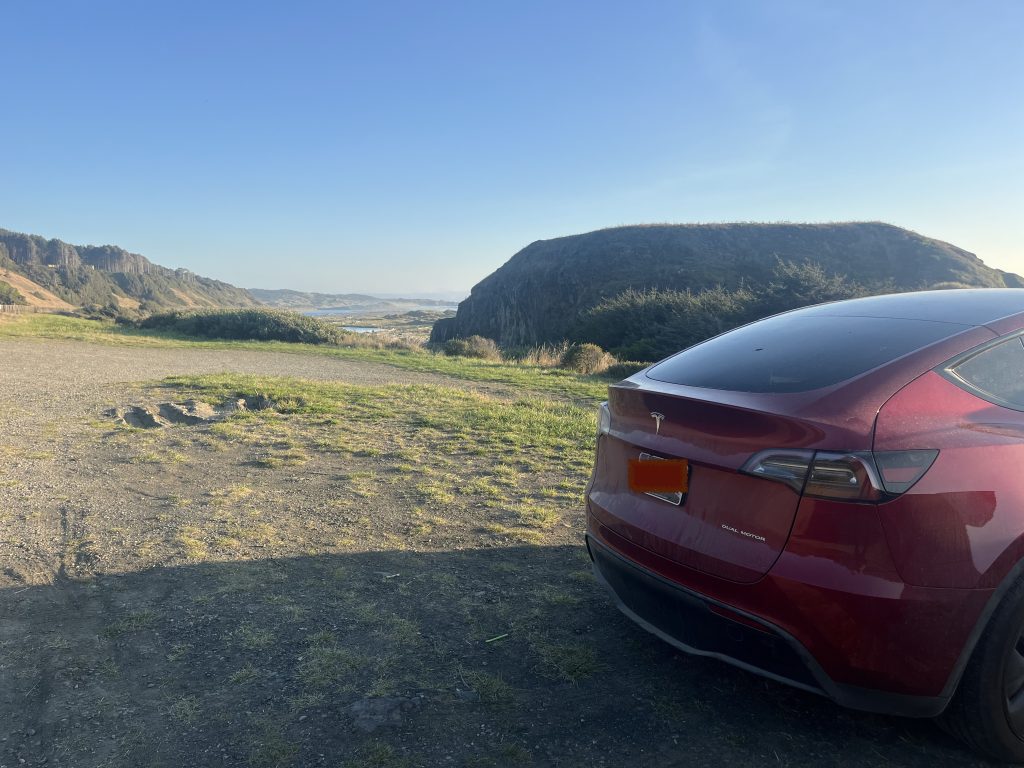 Rear view of a red Tesla parked on a coastal overlook with scenic hills and the ocean in the background