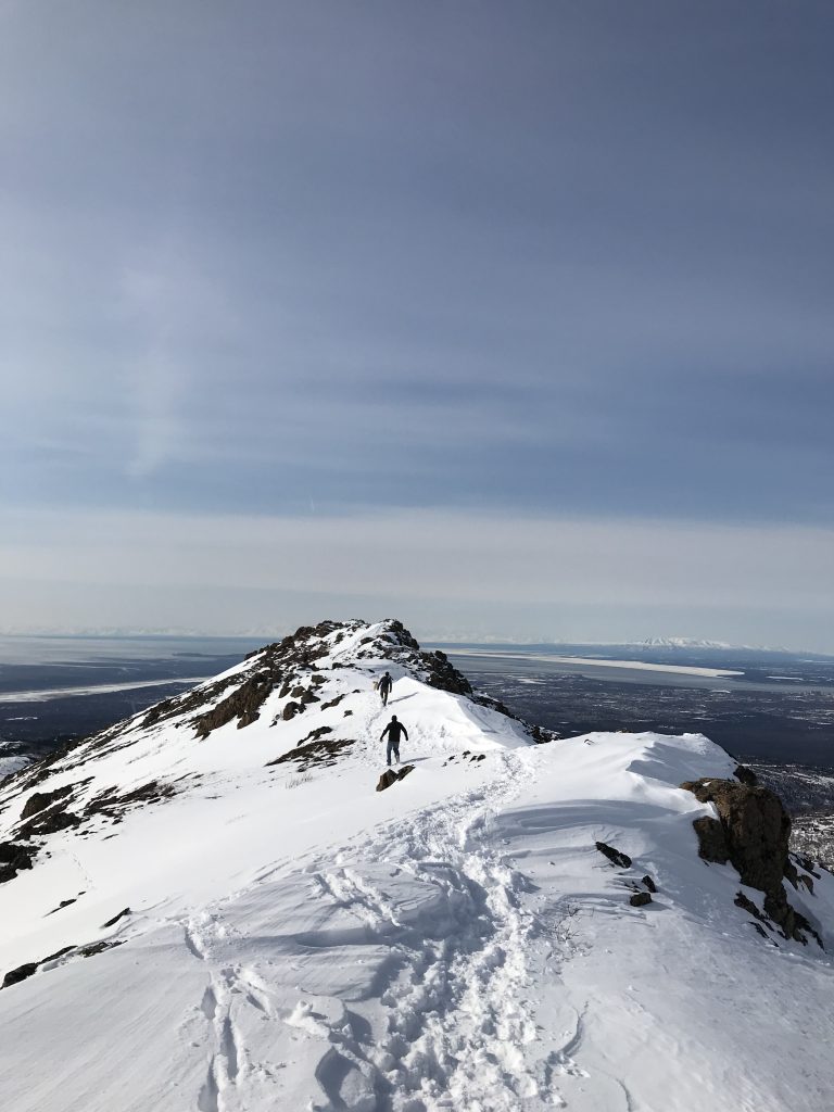 Snow-covered mountain ridge with two hikers walking along the path under a clear blue sky