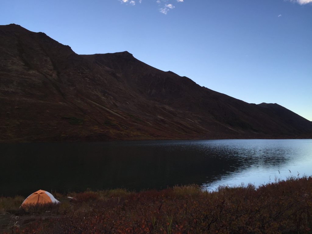 Orange tent set up near the shore of Symphony Lake in the evening, with calm water reflecting the mountains under a clear sky