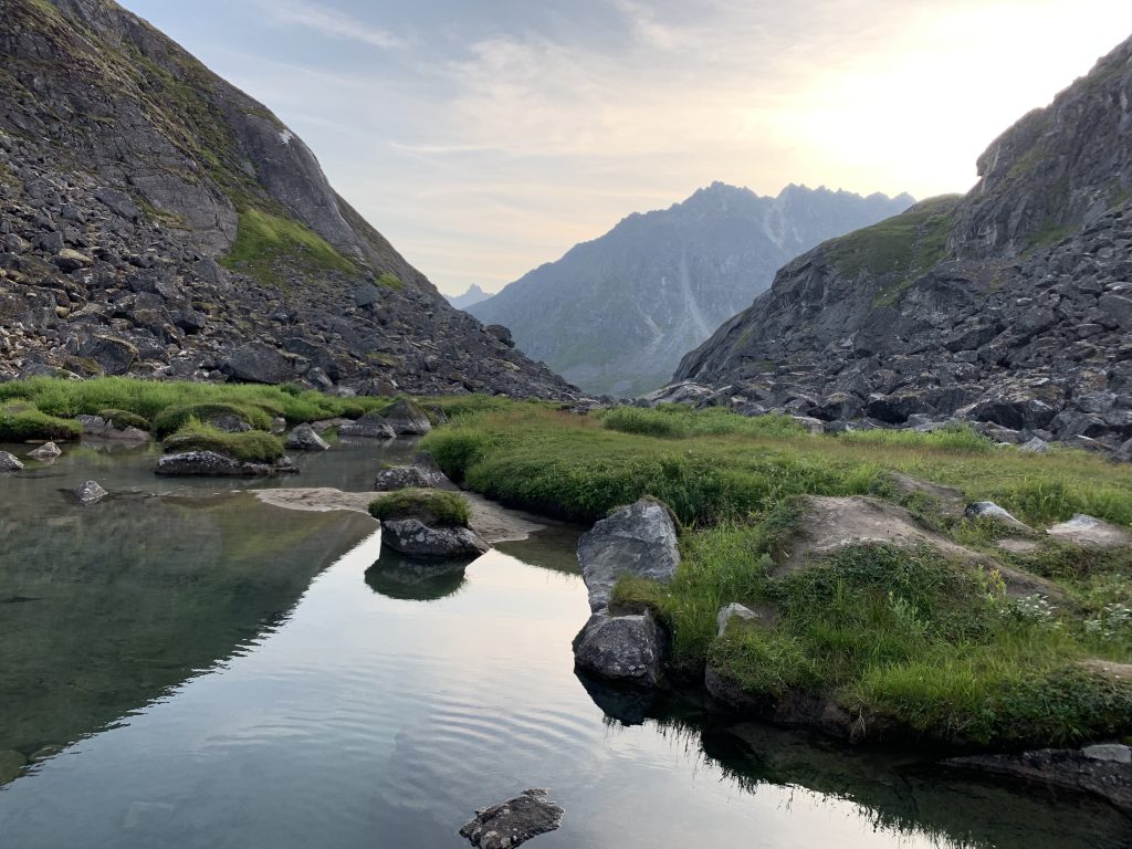 A clear pool reflecting the evening sky, surrounded by rugged cliffs and patches of green grass