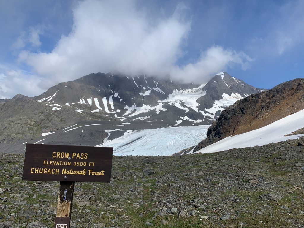 Crow Pass elevation sign with snow-covered mountains and glaciers of Chugach National Forest in the background.