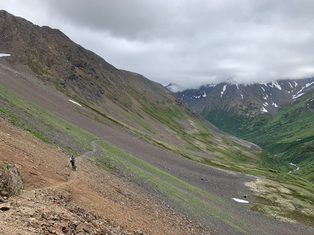 Descending path on Crow Pass into a green valley, with rocky mountains and snow patches under low-hanging clouds.