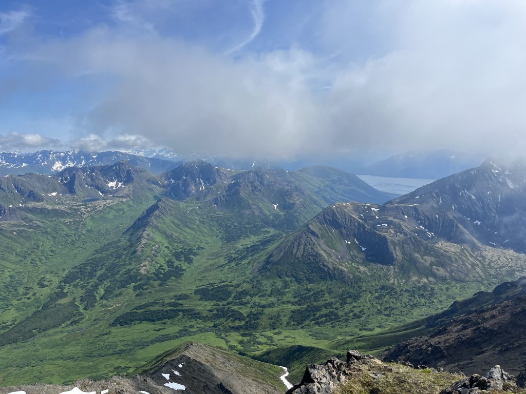 Expansive view of green valleys and mountain peaks from Williwaw Peak, with distant snowy mountains and a light mist drifting over the landscape.