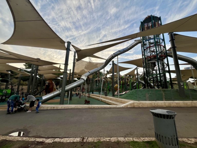 A modern playground in Israel with tall slides, climbing structures, and shaded areas, surrounded by families enjoying the park.