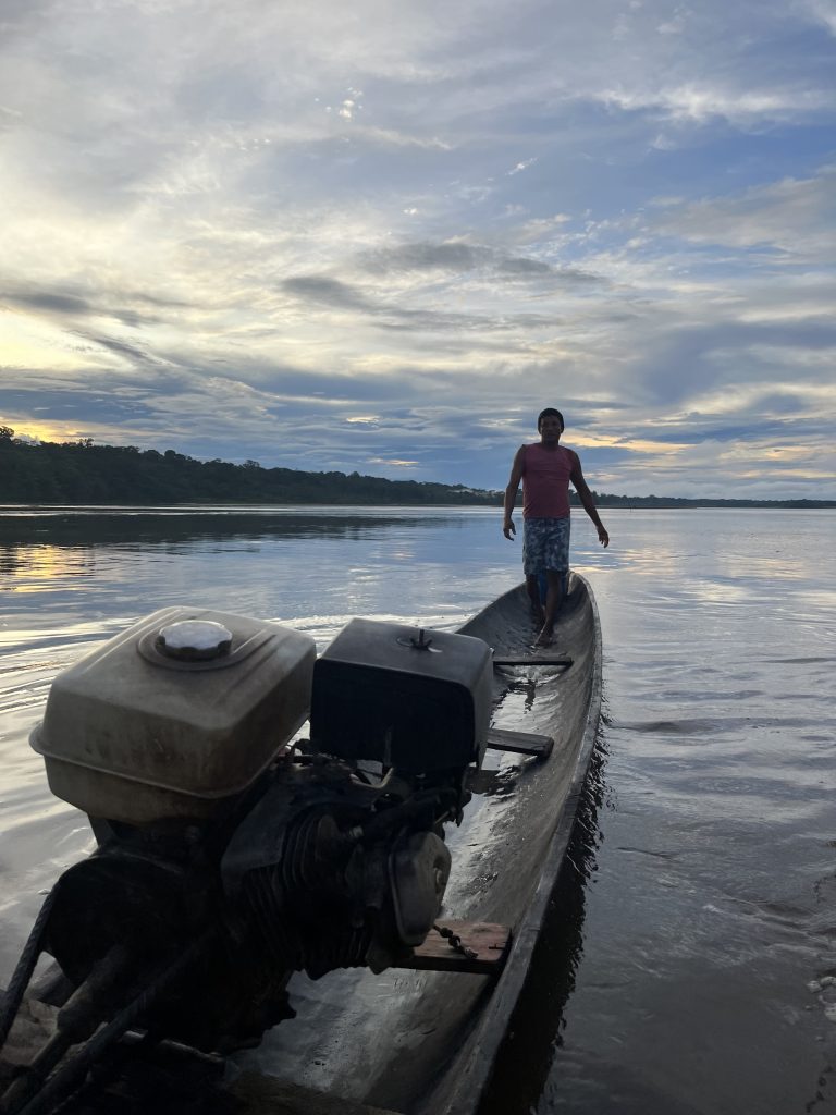 Local man standing on a motorized canoe on the Napo River at sunset, with a wide, calm river behind him