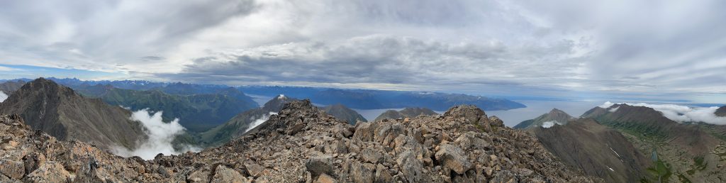 Panoramic view from the summit of South Suicide Peak, showing rocky terrain, mountain ridges, valleys, and distant bodies of water with clouds drifting between the peaks.