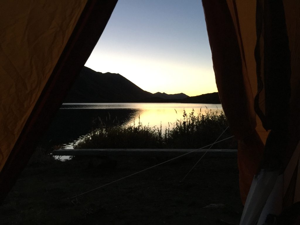 View from inside a tent overlooking Symphony Lake at dusk, with mountains silhouetted against the sky and the lake reflecting the golden sunset