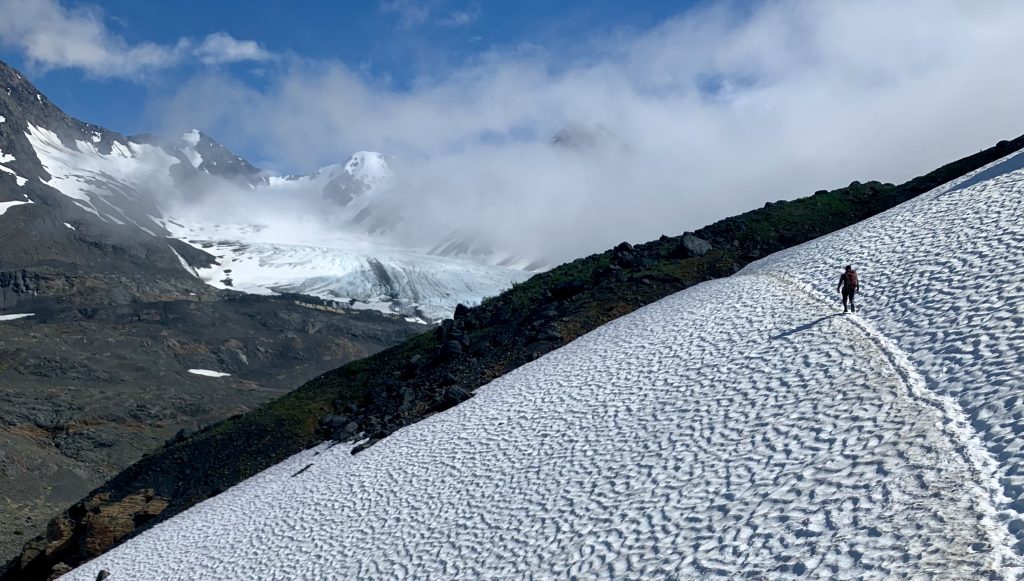 A hiker walking across a snow-covered slope with distant mountains and a glacier in the background