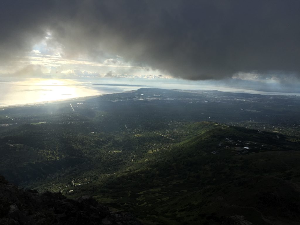 View from Flattop Mountain summit in Anchorage, Alaska, overlooking a vast green landscape, coastal waters, and a dramatic cloudy sky