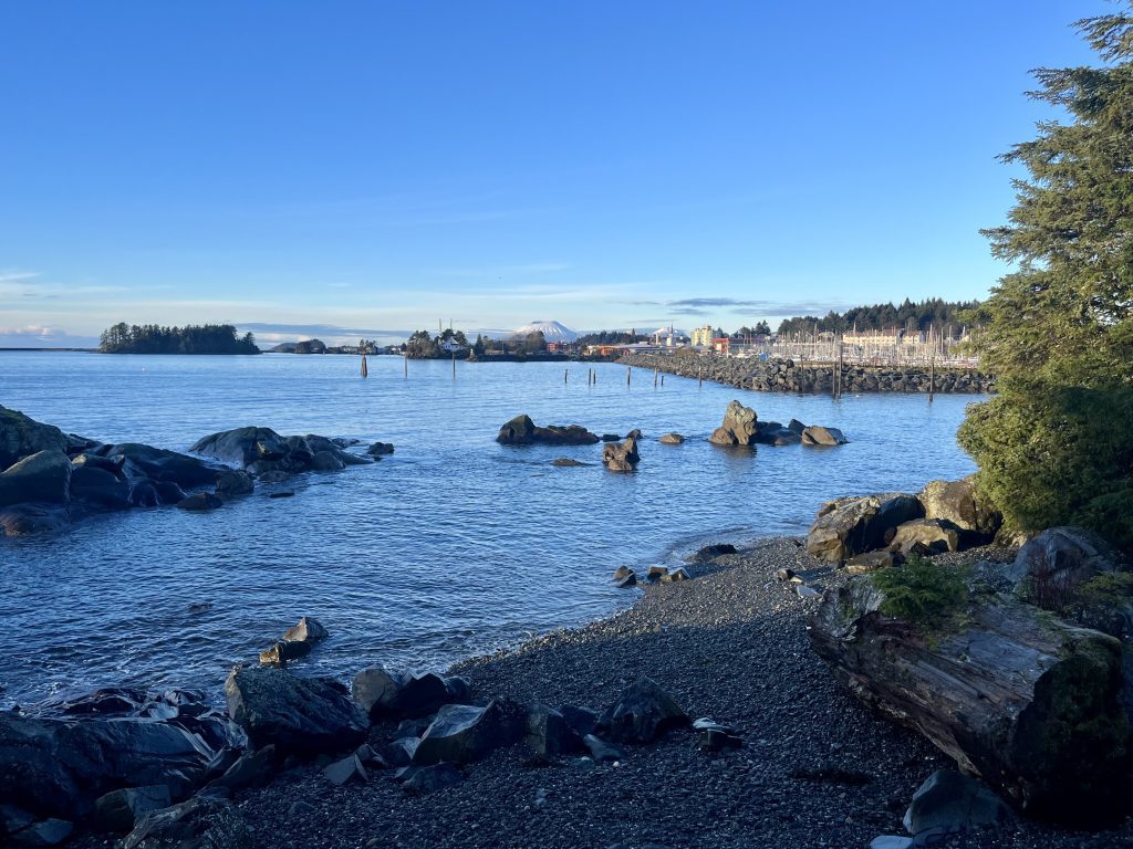 A rocky shoreline with calm blue waters and Sitka town visible in the distance.