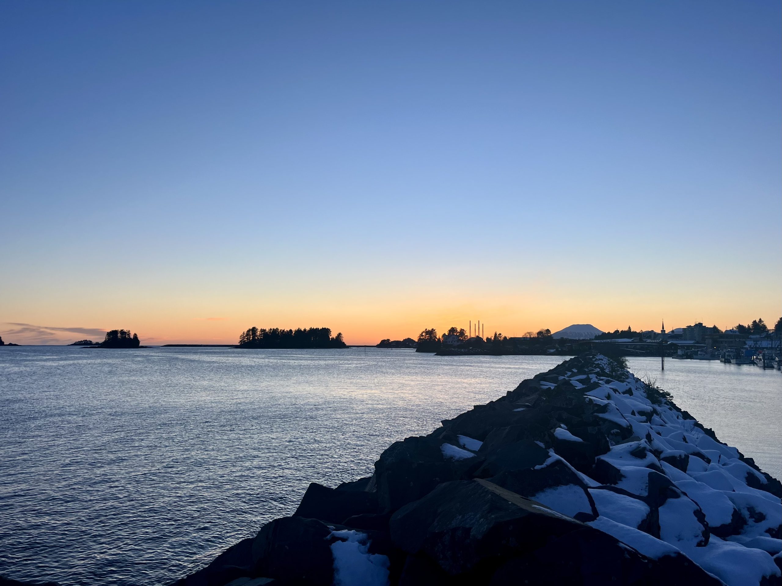 A calm harbor at sunset with small forested islands, a snowy breakwater, and a colorful sky in Sitka, Alaska.