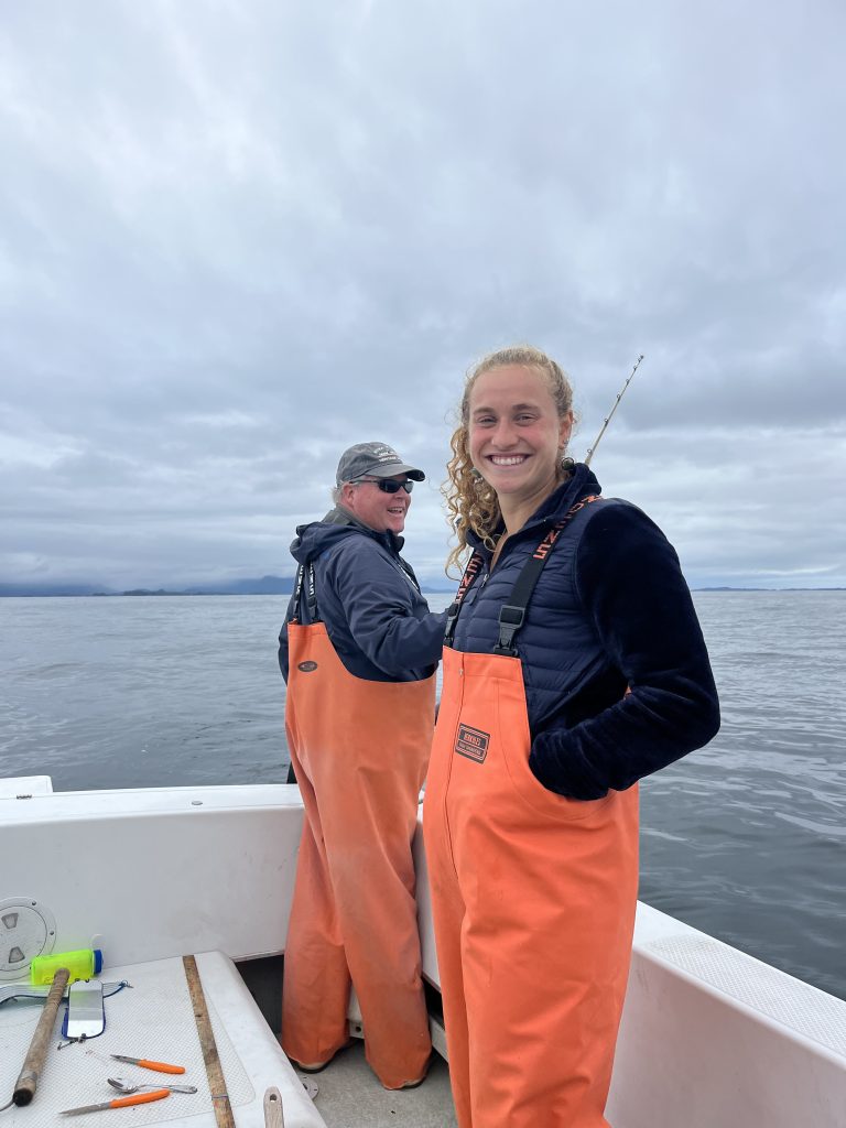 Leah and Tom smiling on a fishing boat in Sitka, Alaska, wearing bright orange fishing overalls on a calm day.