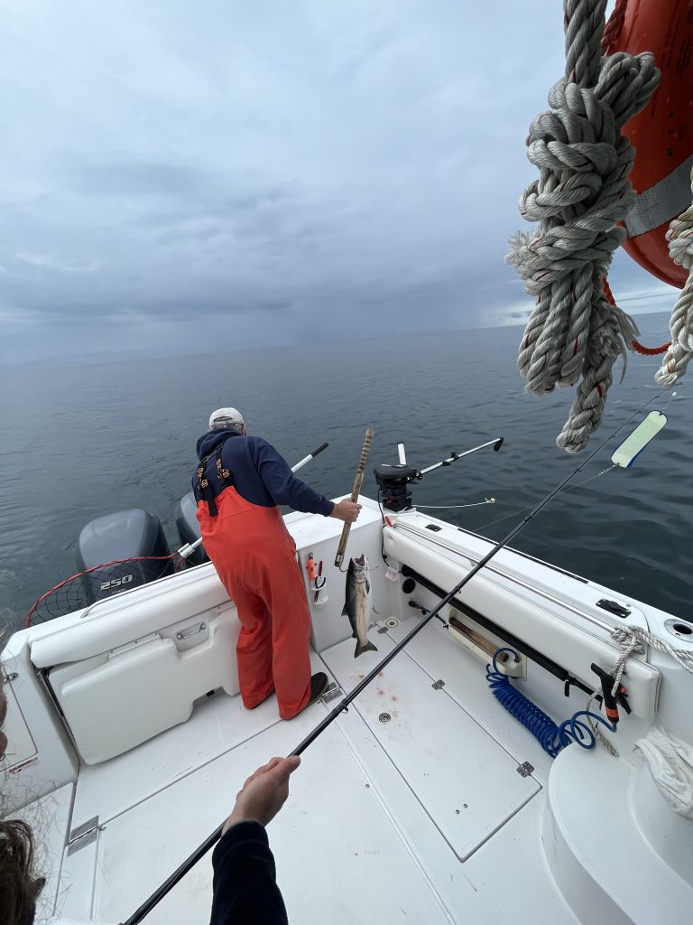 A person in orange overalls holding a fishing gaff near a freshly caught salmon on a charter boat in Sitka, Alaska.